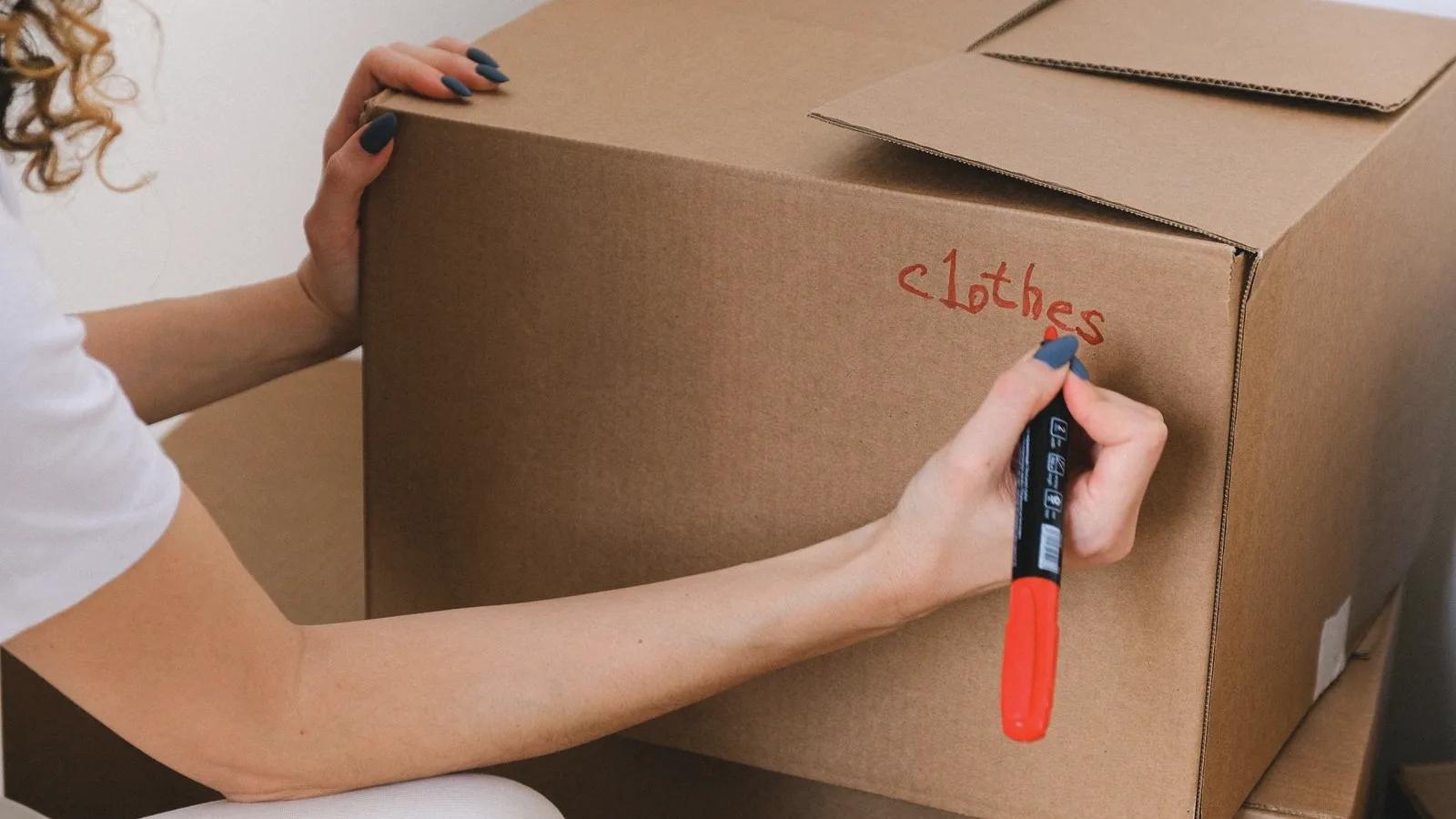 A hand writing a room label on a cardboard moving box with a red marker